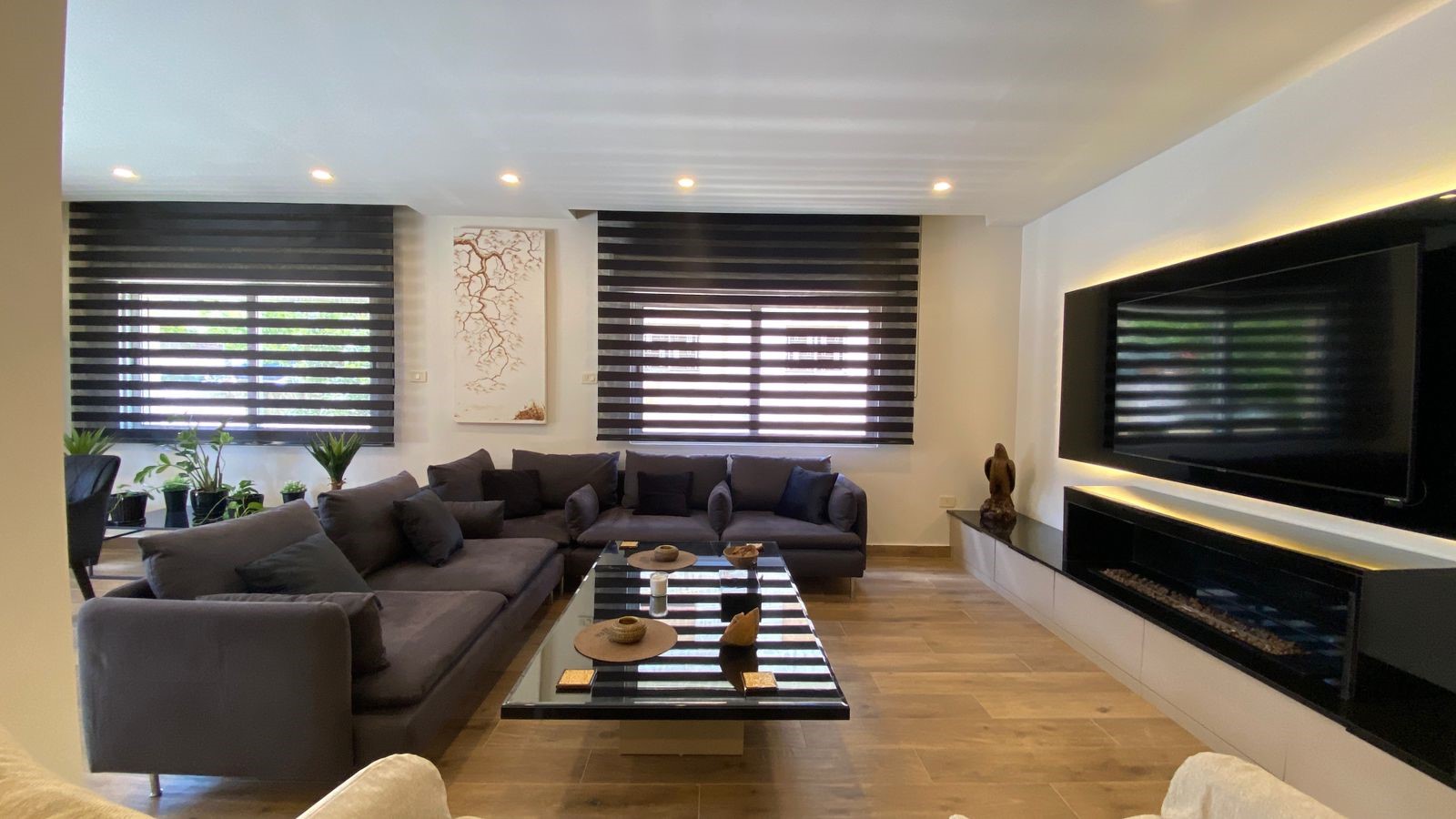 5aeb3406-7cc7-411c-a0e3-e17575bd1c3b Modern living room with a dark gray sectional sofa, striped blackout blinds, and a glass coffee table near a wall-mounted TV.