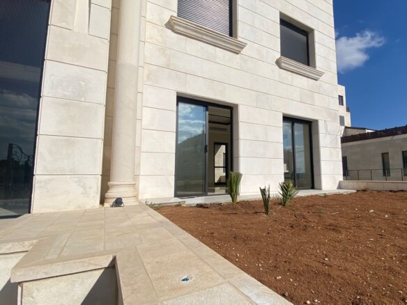 Modern beige stone building entrance with tall glass doors and a decorative column, landscaped with a dirt bed and small plants in front.