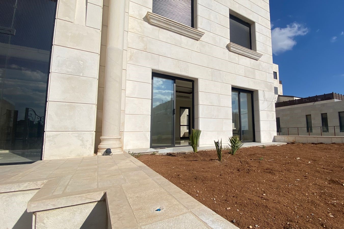5bf3d4ce-c534-4c82-8c14-b4741c1a8444 Modern beige stone building entrance with tall glass doors and a decorative column, landscaped with a dirt bed and small plants in front.