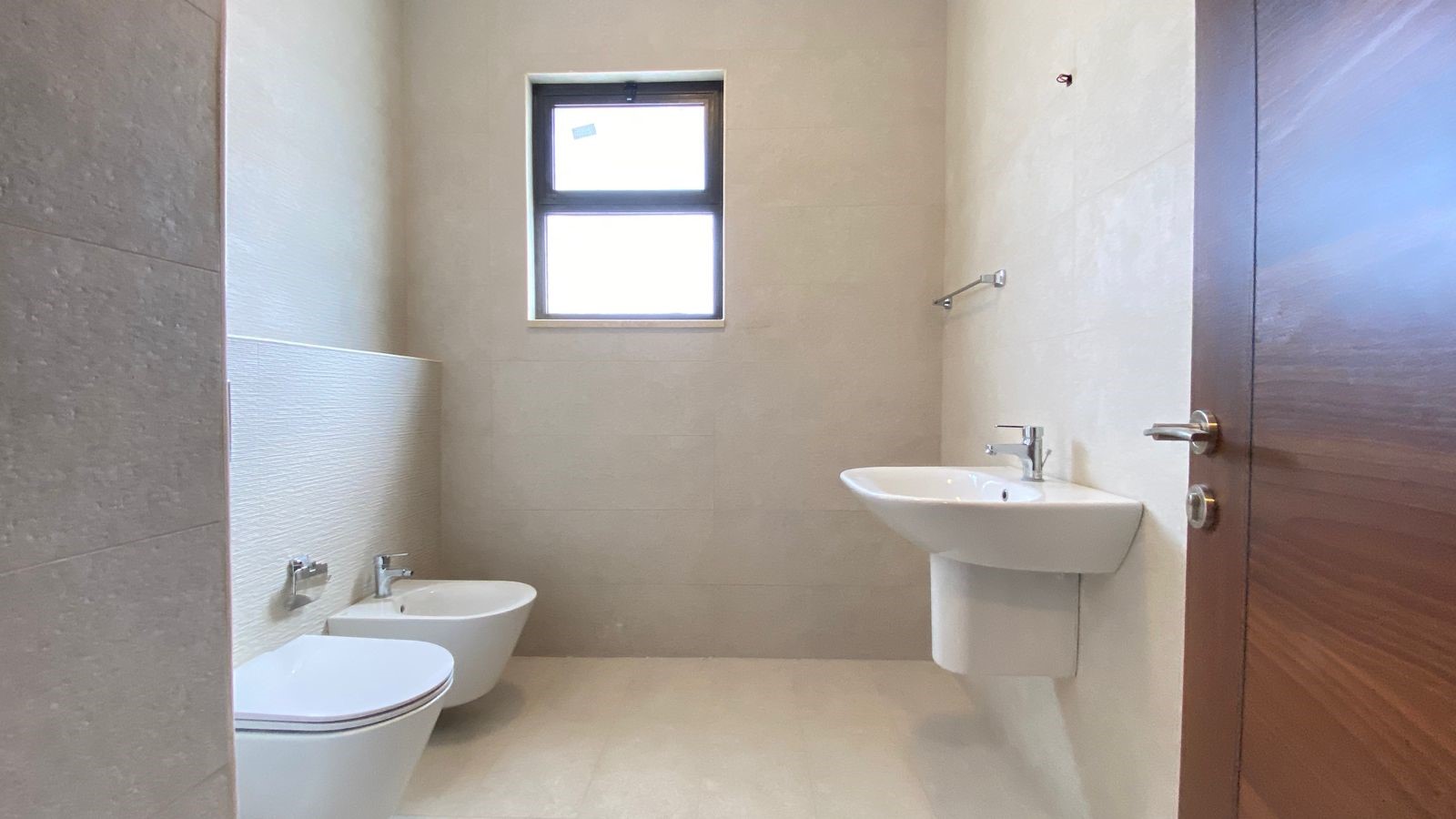 5de31792-8223-4483-82a3-4ff7475e4079 Modern bathroom with wall-mounted sink, toilet, and bidet, beige tile walls, and a small window above.
