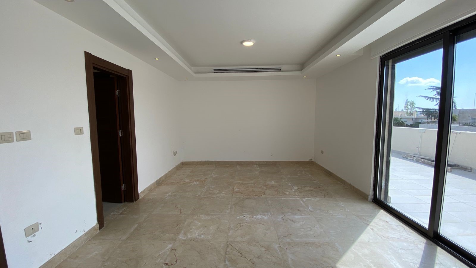 5e2789b7-523c-4695-a8b6-fdb3e449c0b1 Empty living area with large sliding glass doors to a balcony on the right, white walls, beige marble tile floor, and a recessed ceiling with small lights.