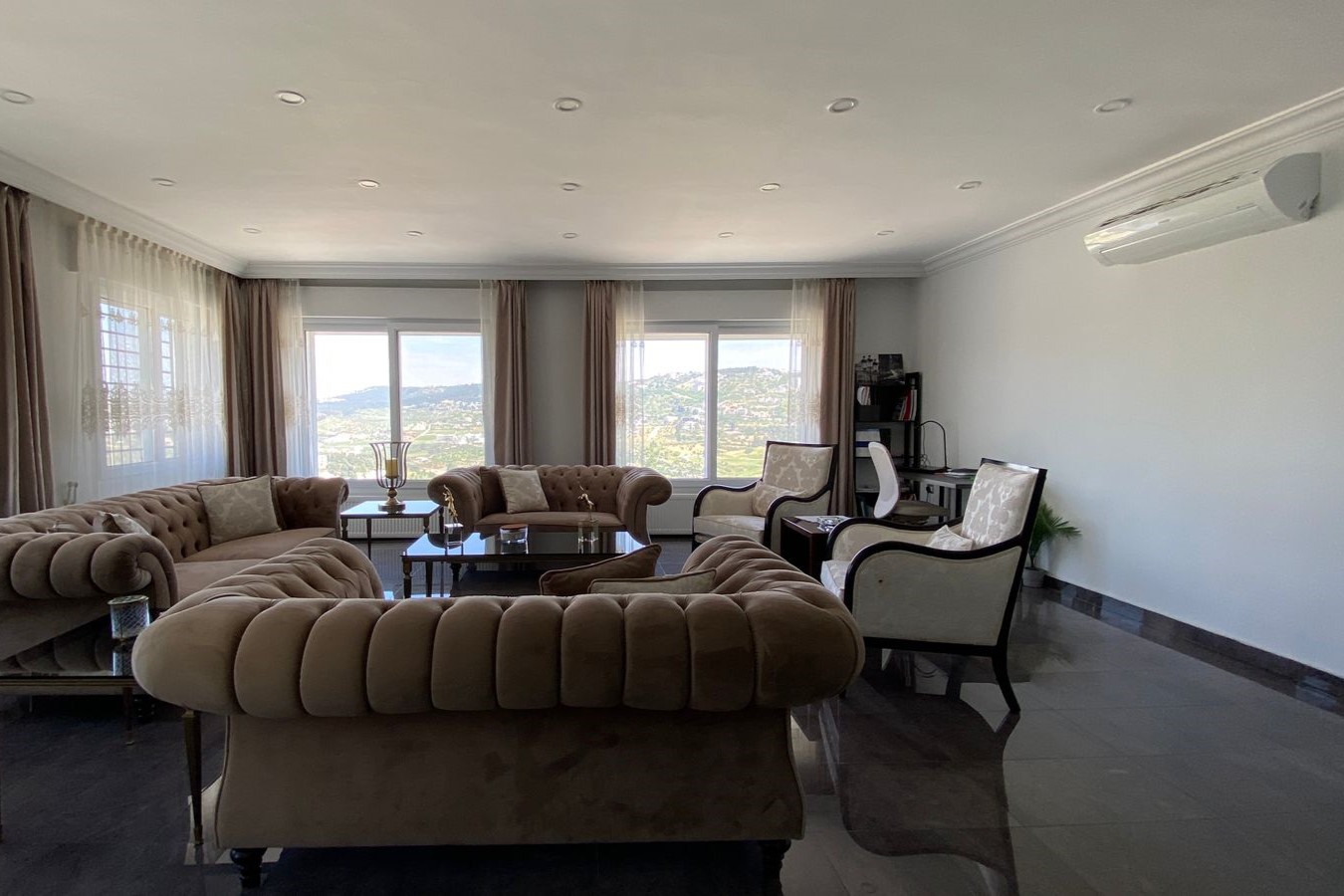 5f3b6170-a06a-4acd-a133-a62e9447bf8c Spacious living room with beige tufted sofas, glass coffee tables, and large windows with sheer and brown curtains overlooking hills.