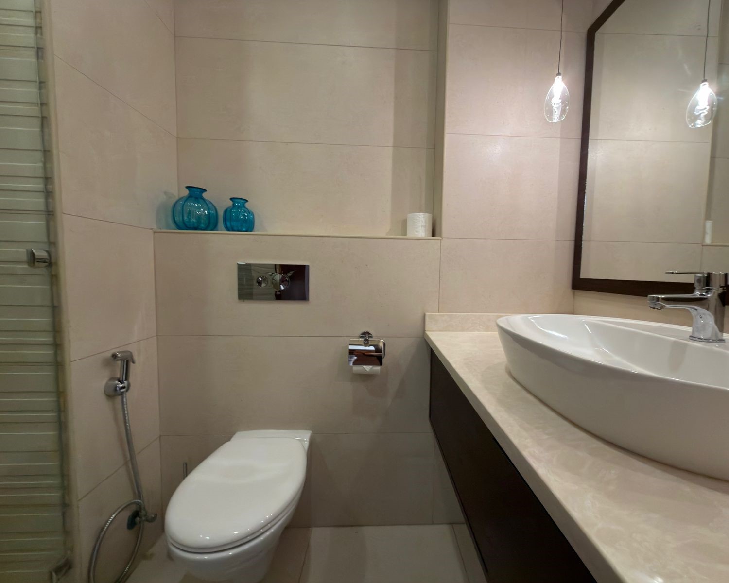 604b3edc-443c-4bc3-9c44-429647e5301c Modern bathroom with cream tiles, a white countertop sink, and a wall-mounted chrome faucet; a dual blue glass vases display sits on a recessed shelf above the toilet area.