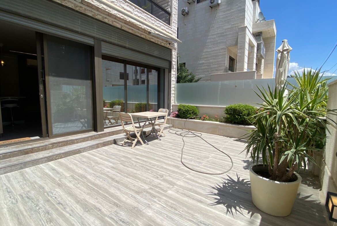 Patio area with sliding glass doors, a small table and two chairs on a light wooden deck, hose on the ground nearby.