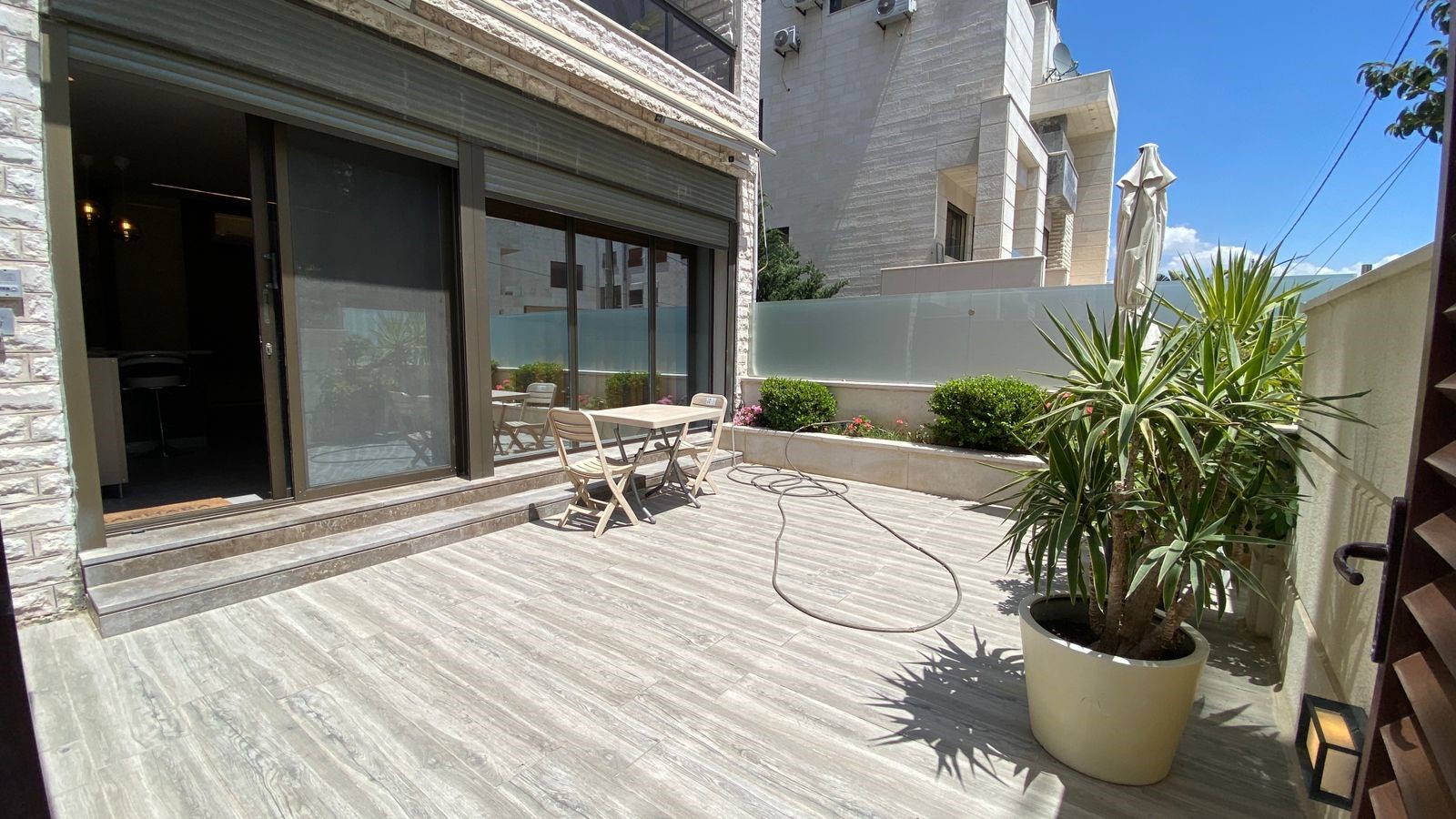 61507b07-f447-49b0-aec3-47641f0eee06 Patio area with sliding glass doors, a small table and two chairs on a light wooden deck, hose on the ground nearby.