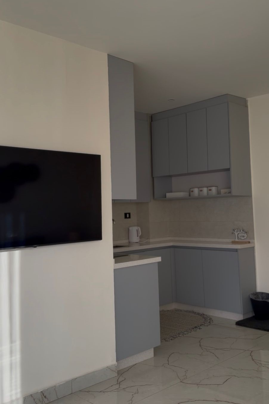 61523ffa-a0d0-4035-9cf6-b58e134dbdd6 Flat-screen TV mounted on a white wall overlooks a modern gray-kitchen with upper and lower cabinets and a marble floor