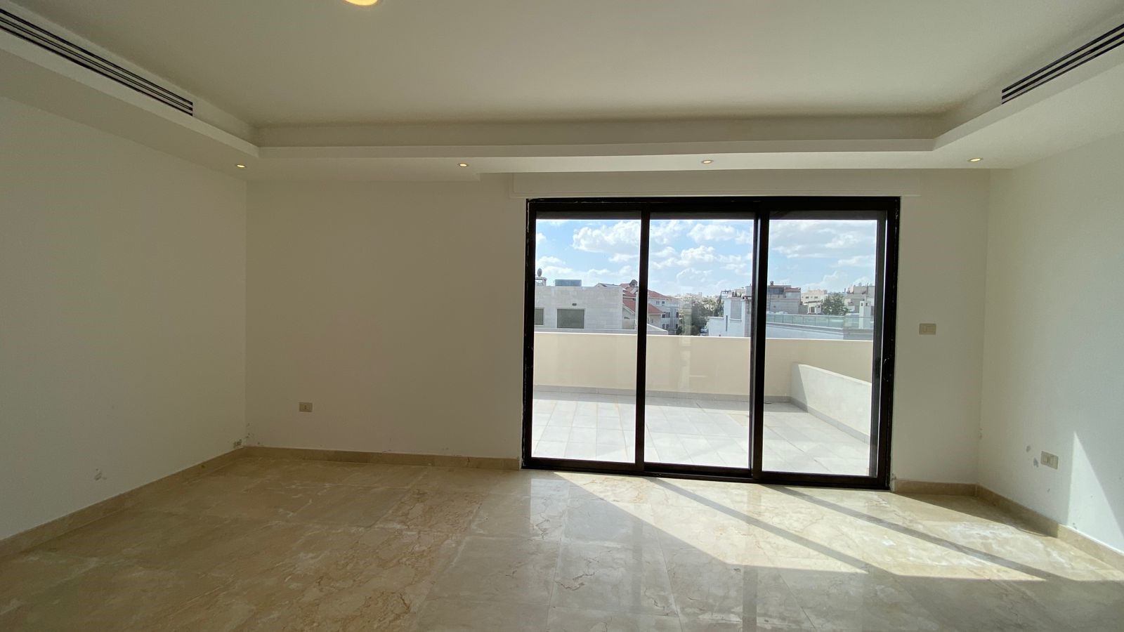 61553f4e-7e0e-4710-b0fc-2eb35397d5dd Empty living room with beige marble floor, white walls, and large sliding glass doors opening to a sunlit balcony with a city view.