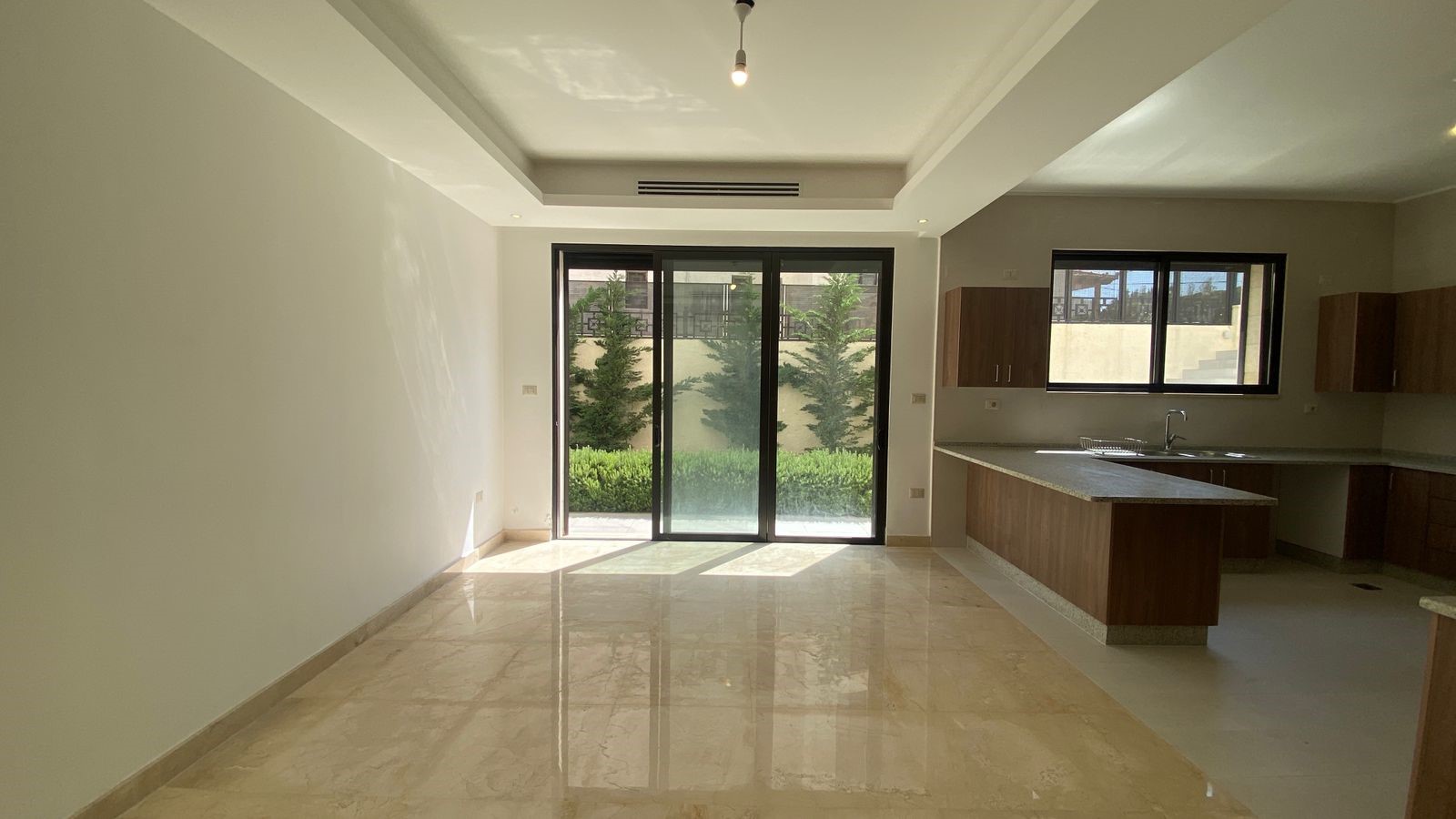 6162c2c4-ba03-4100-9f0d-b1f884f02b73 Bright empty open-plan living area with marble floors, large sliding glass doors to a green backyard, and a modern kitchen on the right.