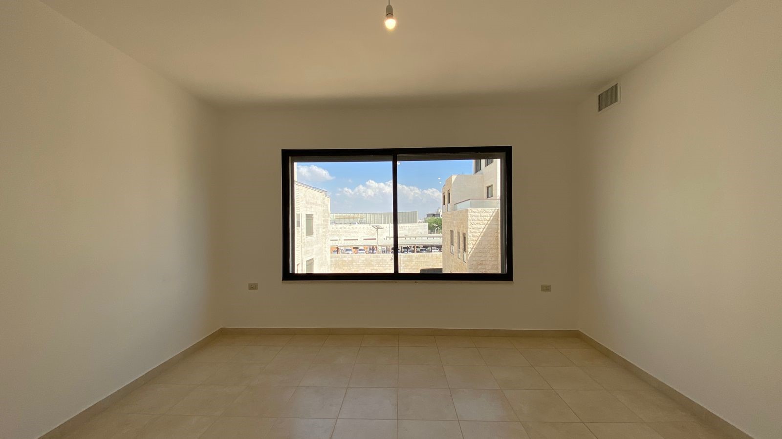 61be6d26-04f6-434c-80c3-93f7ab271504 Empty, unfurnished room with tiled floor and a large window; outside are beige brick buildings and a blue sky.