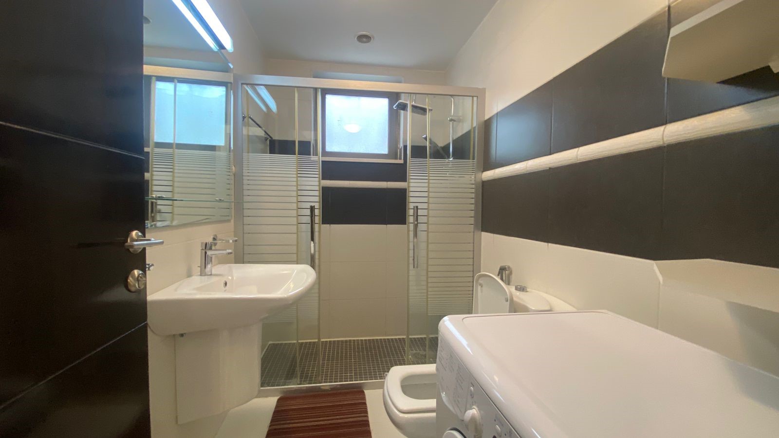 61d0459d-eb56-4148-8723-ae90fa7c6dff Modern bathroom with glass shower enclosure, white sink, toilet, and a front-loading washing machine; black and white tiled walls create a contemporary look.