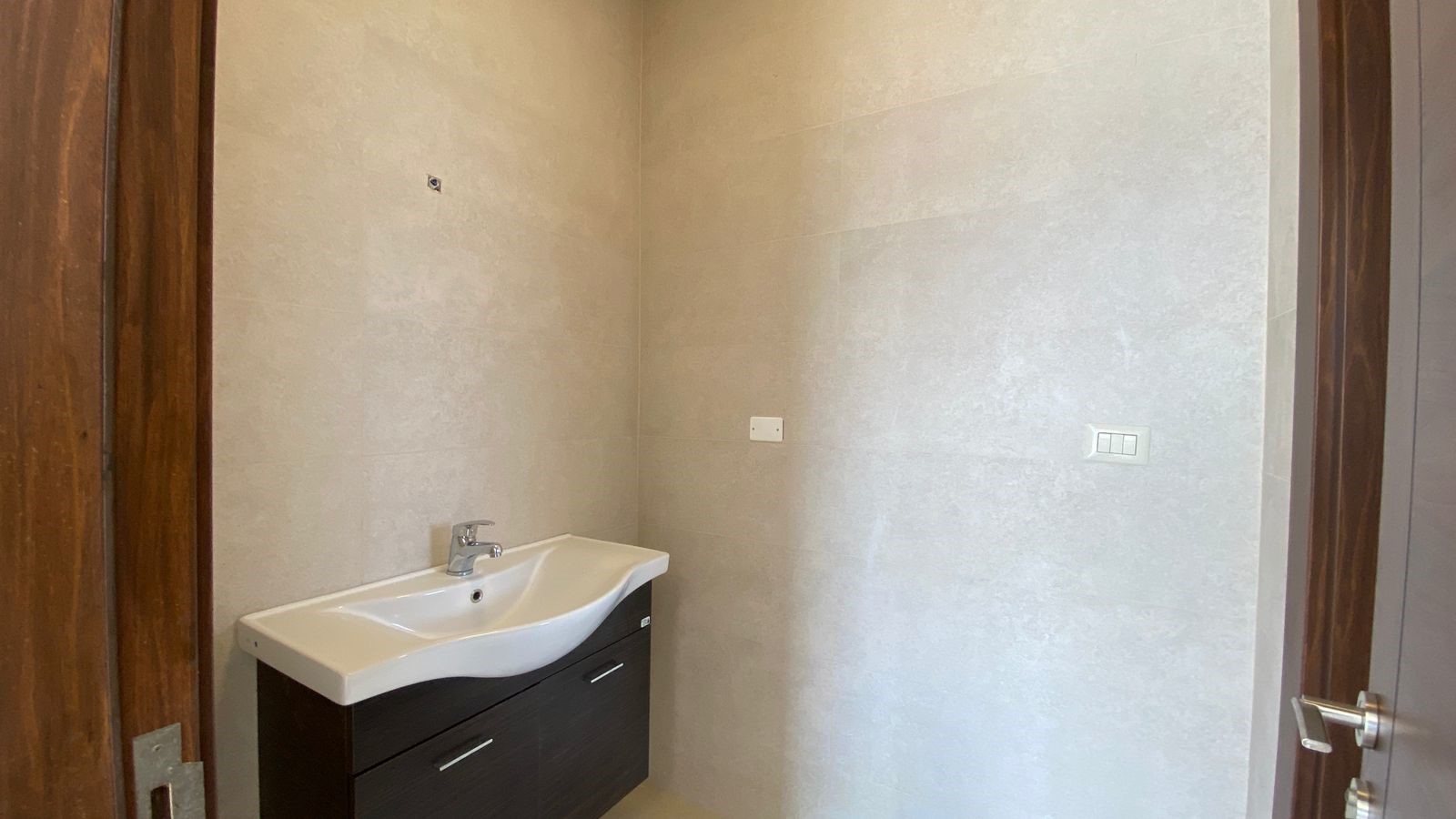 63ec49ff-373e-4513-8c07-bbd4264b098c Small bathroom with a white sink and dark wood vanity against beige tiled walls; door frames visible on both sides.
