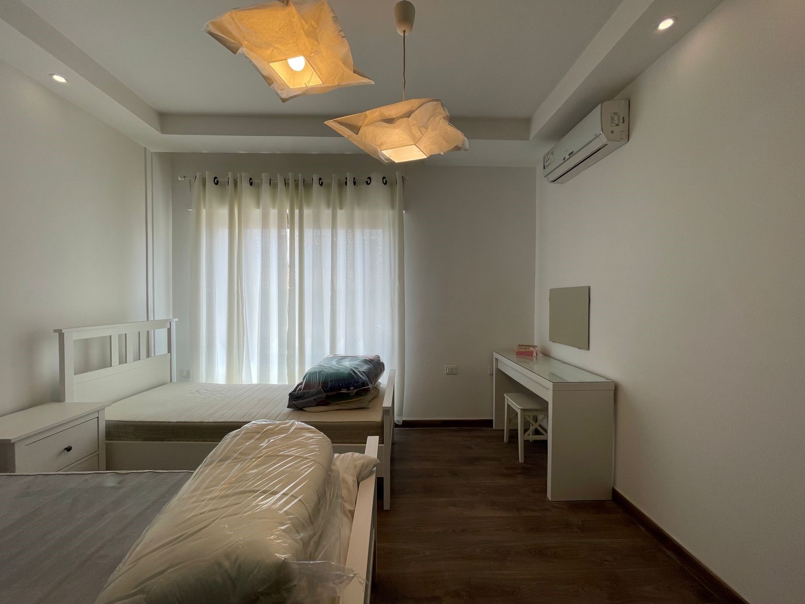 64fd5c3d-3b6b-4c16-be4b-be0a9e8805a5 Minimalist white bedroom with two single beds, a desk with stool, and sheer-curtained window; mattresses wrapped in plastic.
