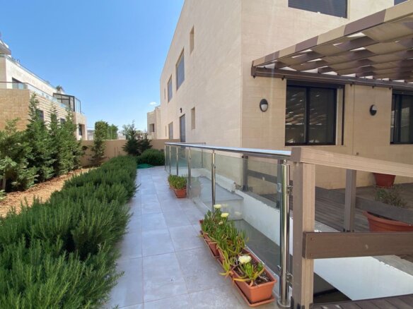 Outdoor walkway between beige stone buildings with a glass railing, potted plants, and neatly trimmed hedges under a clear blue sky.