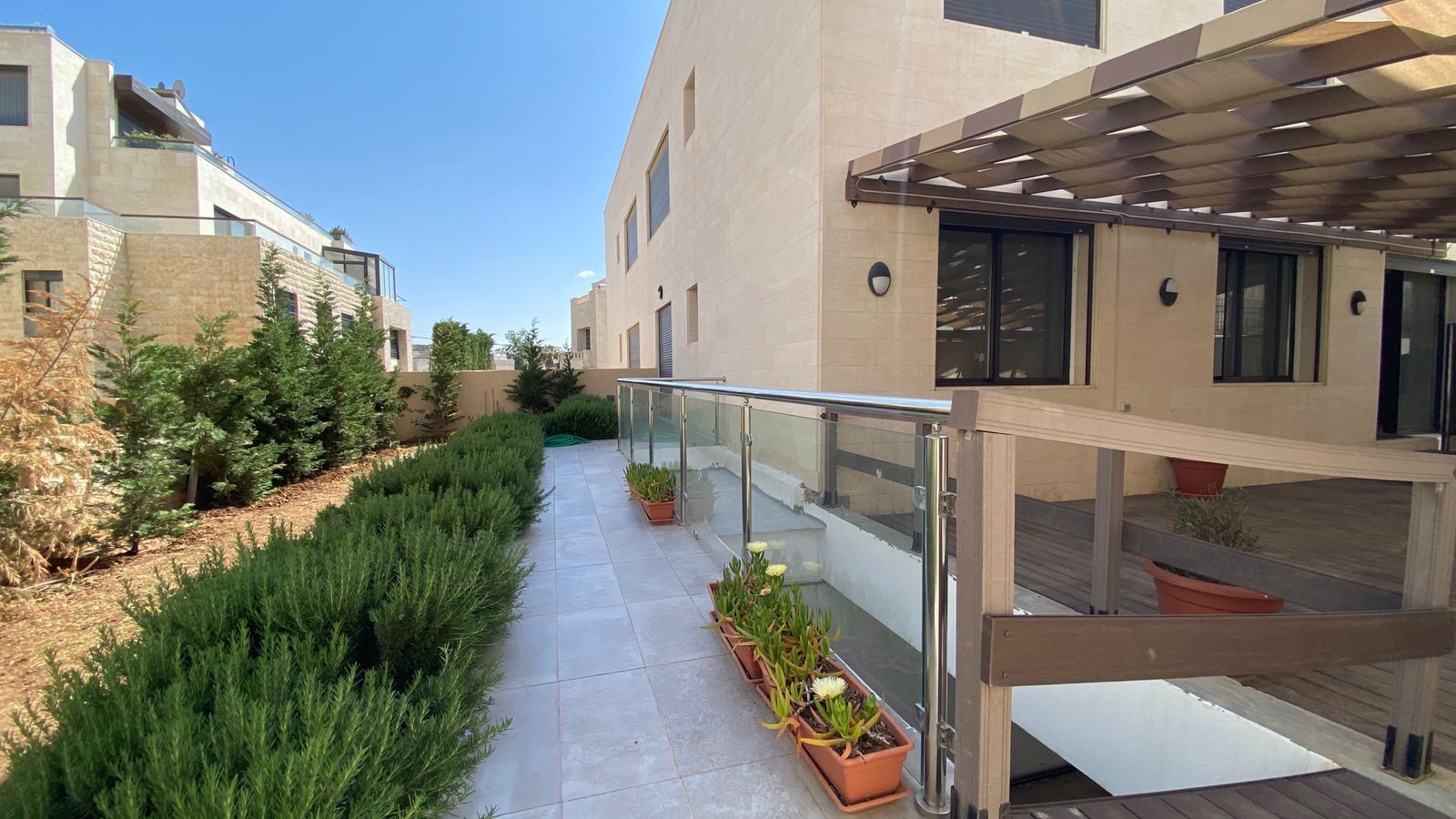 65372c1d-14dc-4f7b-9999-181018c79835 Outdoor walkway between beige stone buildings with a glass railing, potted plants, and neatly trimmed hedges under a clear blue sky.