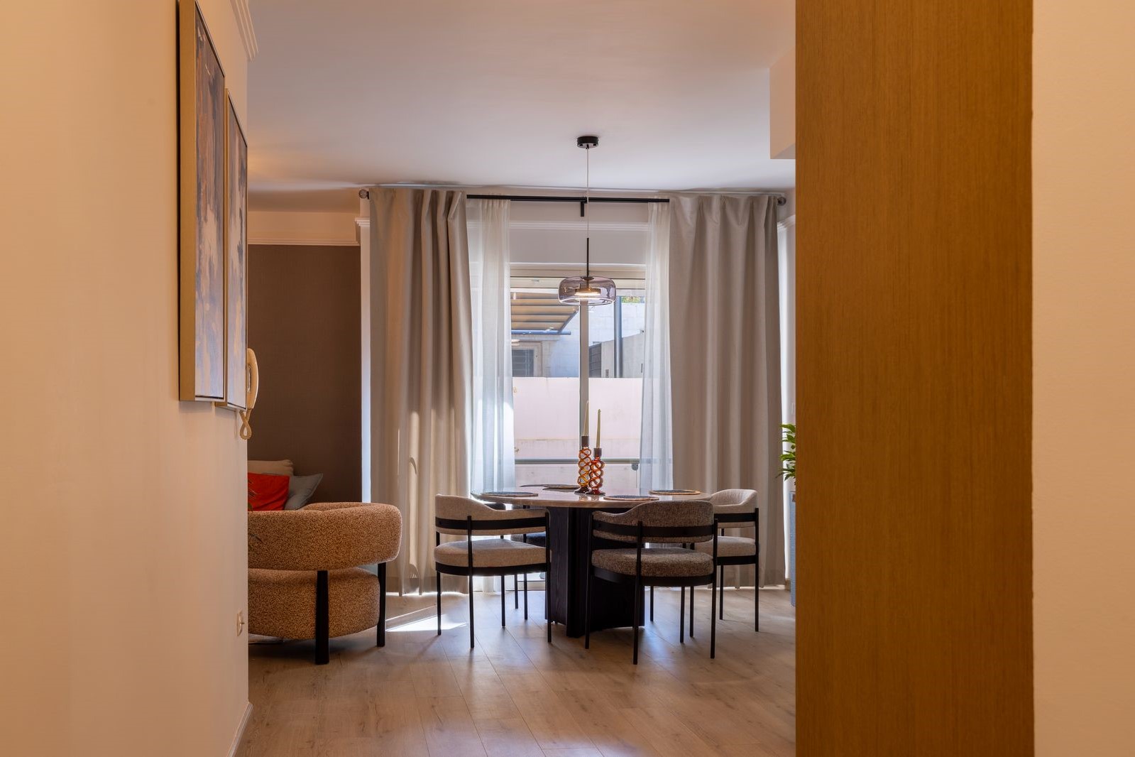 6756001d-6f30-498e-933e-d4fec6f40081 A round dining table with four upholstered chairs, a pendant light, and a candle centerpiece, in a sunny dining nook with sheer curtains.