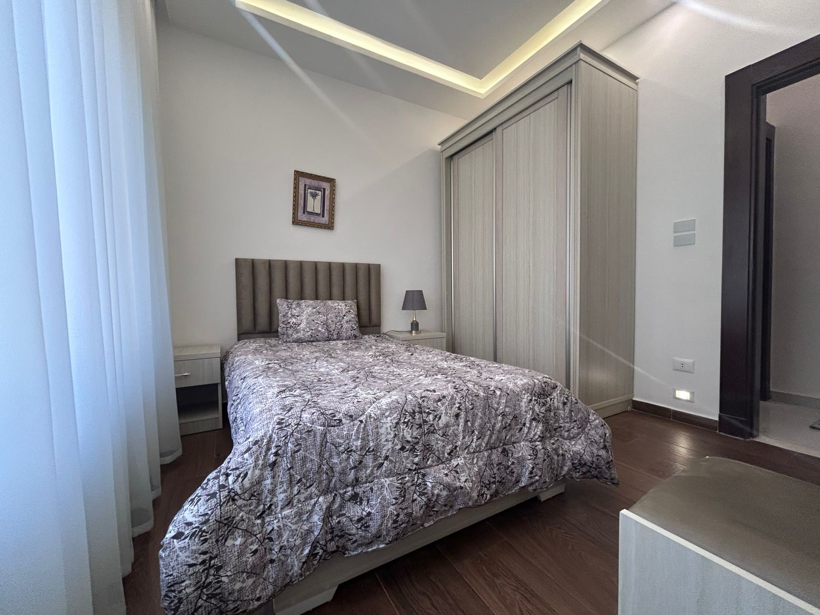 68c49dfd-32c9-4ec7-9093-da6a127e60a4 Modern bedroom with a gray upholstered headboard, patterned quilt, two white nightstands, and a tall sliding-door wardrobe.