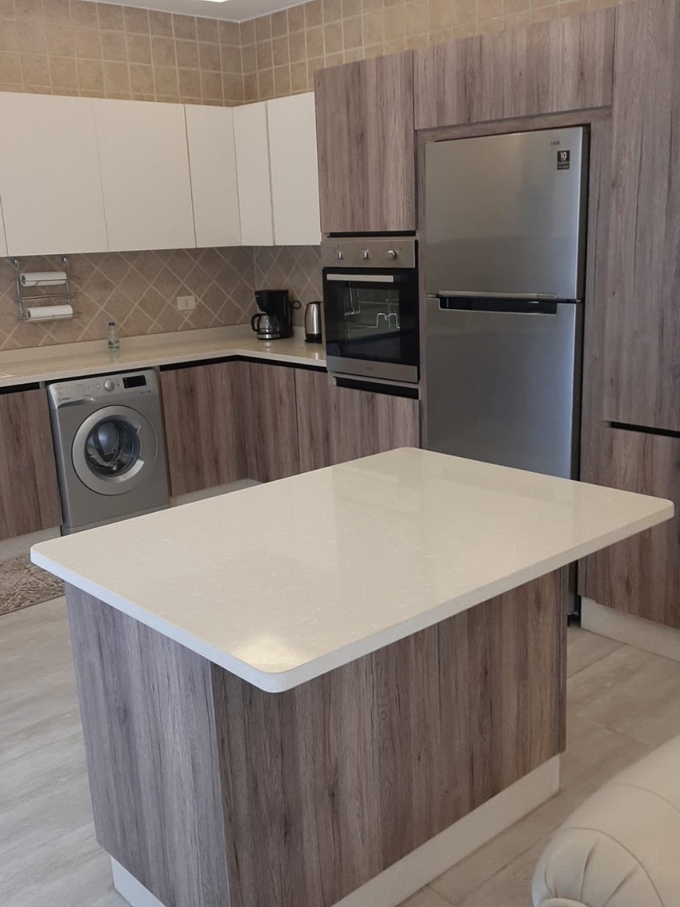 690c590b-0a32-4f16-94ef-1789e67cb543 Modern kitchen with wood veneer cabinets, white countertops, and stainless steel appliances including a fridge and built-in oven; island at center.