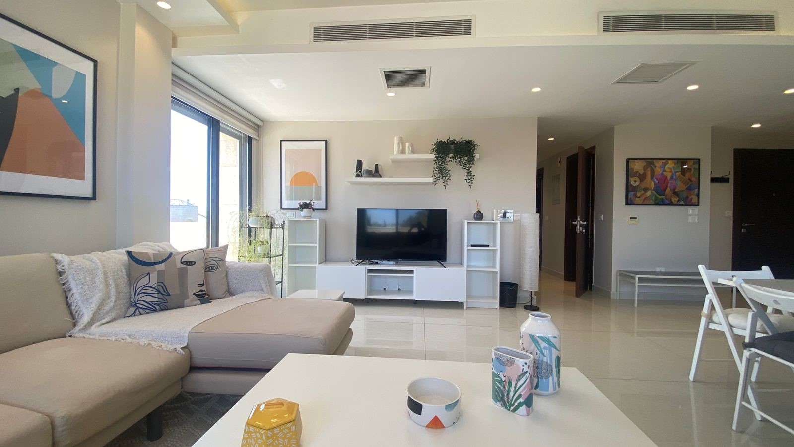 6915f1b9-6f78-4782-ac37-1cec988d6588 Bright contemporary living room with a beige sectional, decorative pillows, and a white coffee table in the foreground; a TV on a white entertainment unit flanked by shelves and plants in the background.