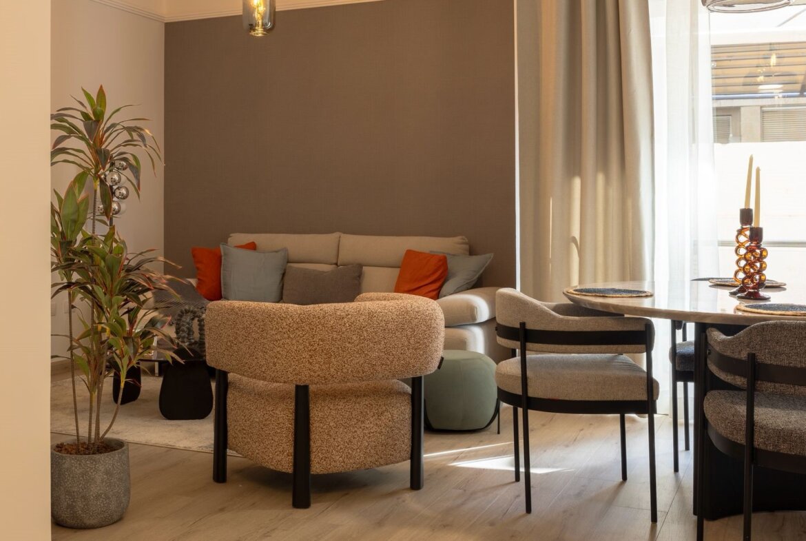 Cozy living area with a beige sofa, orange and gray cushions, and a curved textured ottoman near a planter in a sunlit room.