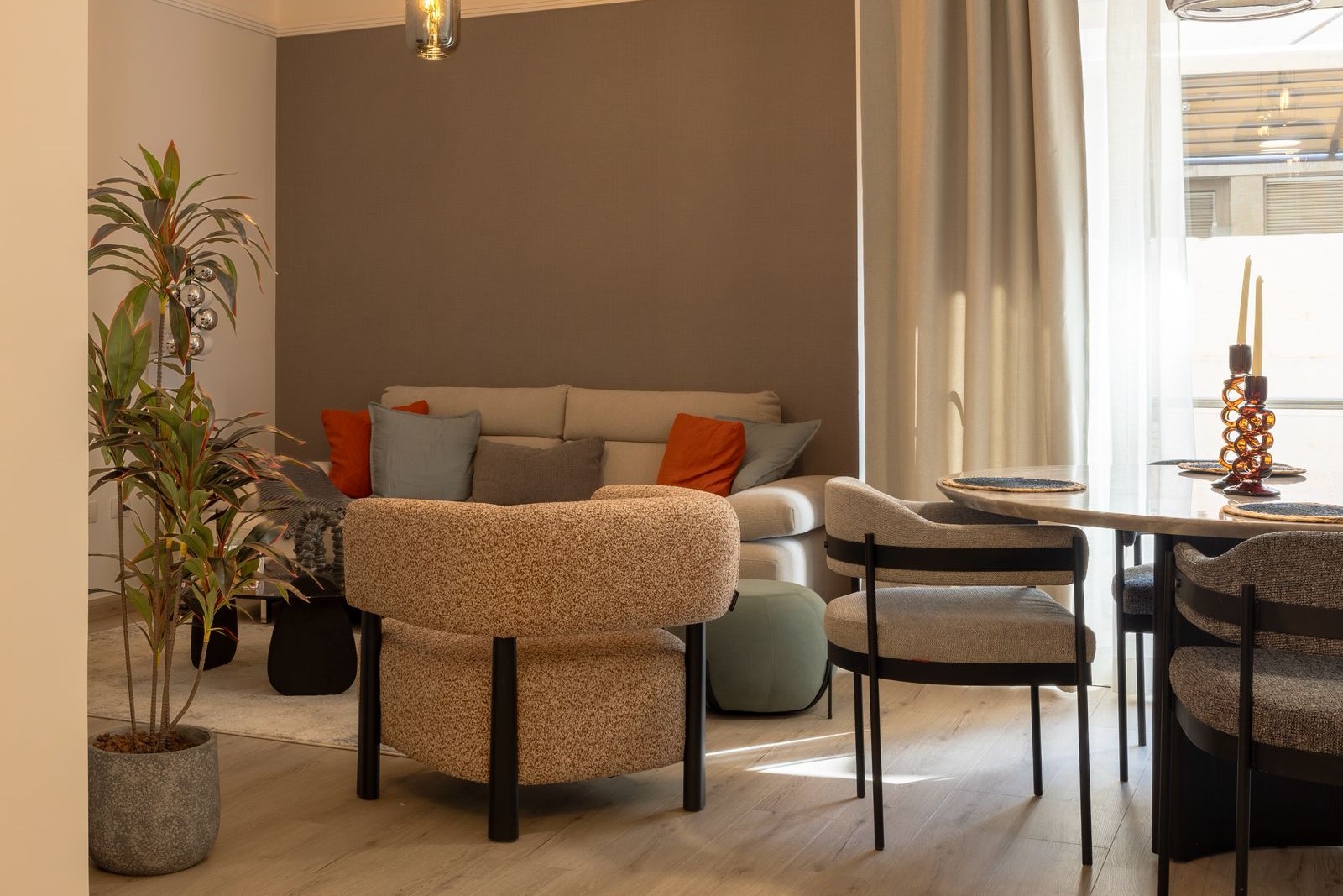 6a57313d-c4bd-407d-89ec-a00fb32280db Cozy living area with a beige sofa, orange and gray cushions, and a curved textured ottoman near a planter in a sunlit room.