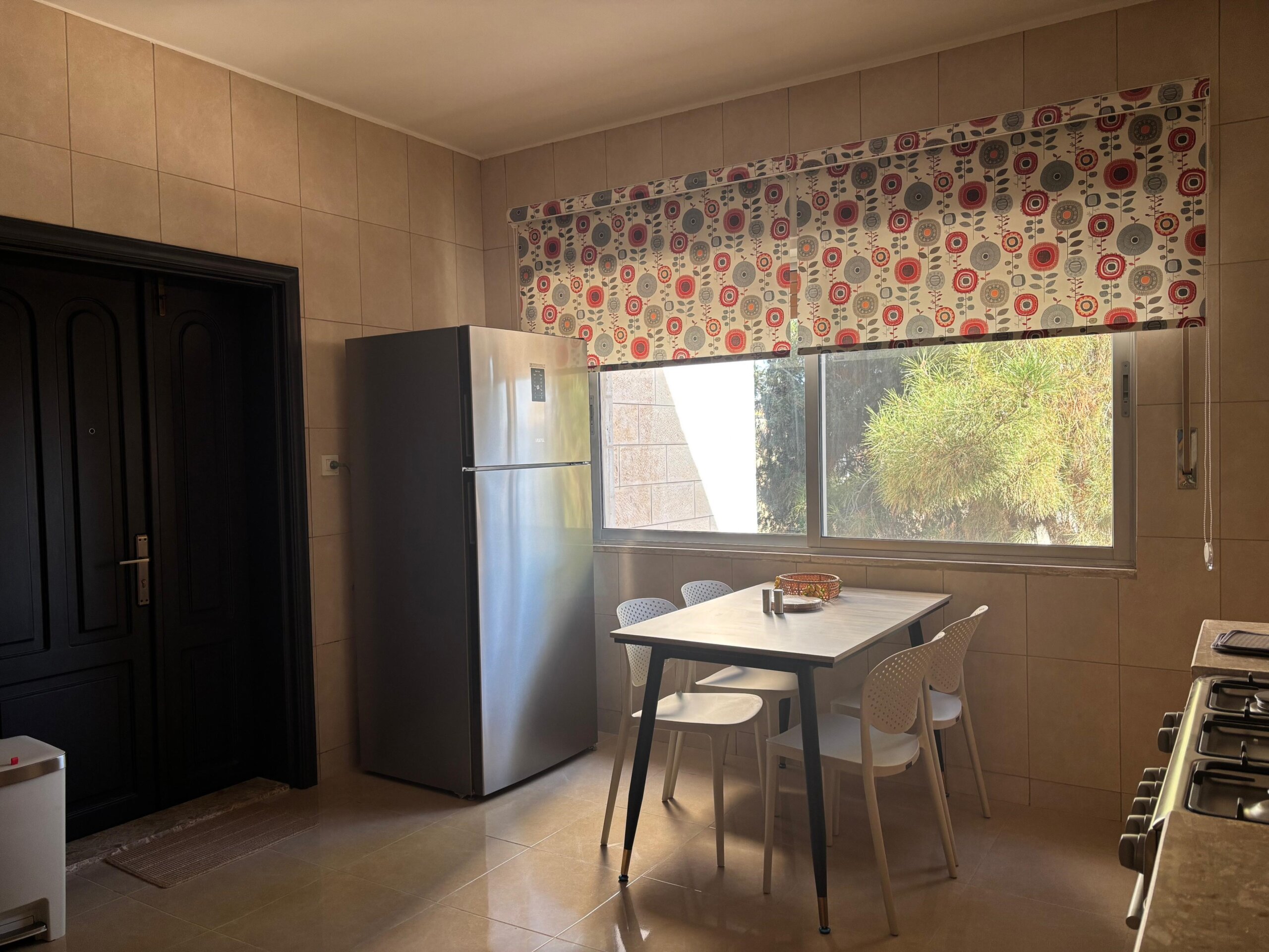 6bb75cd4-d6a7-4fb7-b18a-bf7f1e7d988c Kitchen with a stainless steel fridge beside a large window, a white dining table and four white chairs, patterned roller blinds above the window.