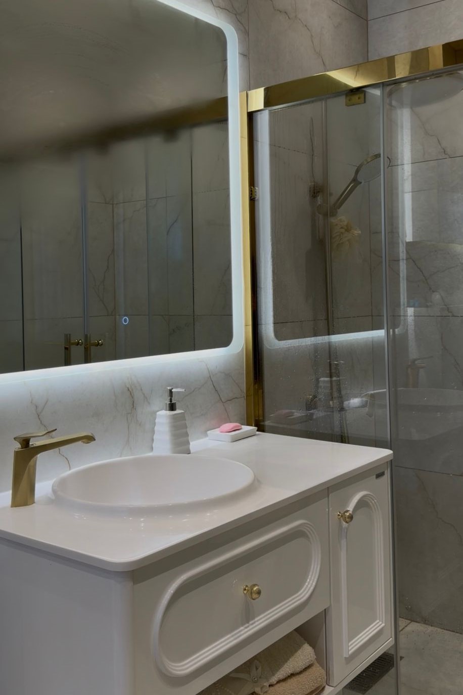 6bc75a8c-e21f-4351-9d0d-517e84127601 White bathroom vanity with an oval sink, gold faucet, soap dispenser, and marble countertop, reflected in a backlit mirror beside a glass shower door with brass trim.