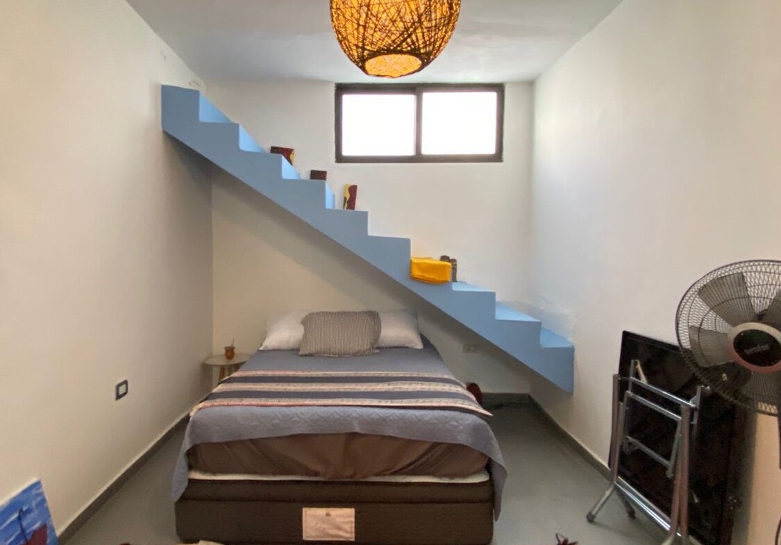 Bedroom with a blue diagonal staircase above a bed, window at the far wall, and a woven pendant light overhead.”,