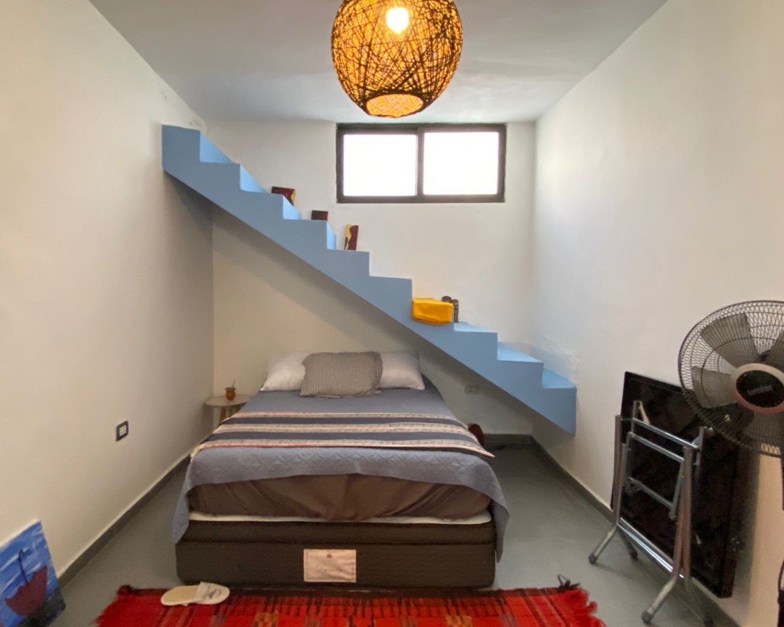 Bedroom with a blue diagonal staircase above a bed, window at the far wall, and a woven pendant light overhead.”,
