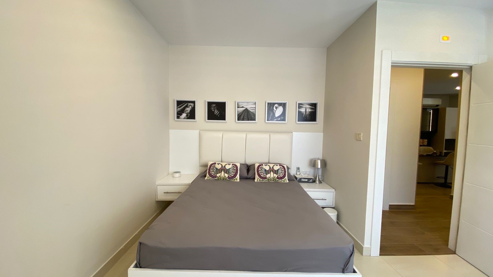 6e841e6a-f271-4f36-a56e-632885dc251f Modern bedroom with a white padded headboard, gray bedspread, and two nightstands flanking a queen bed; black-and-white wall art above the headboard.