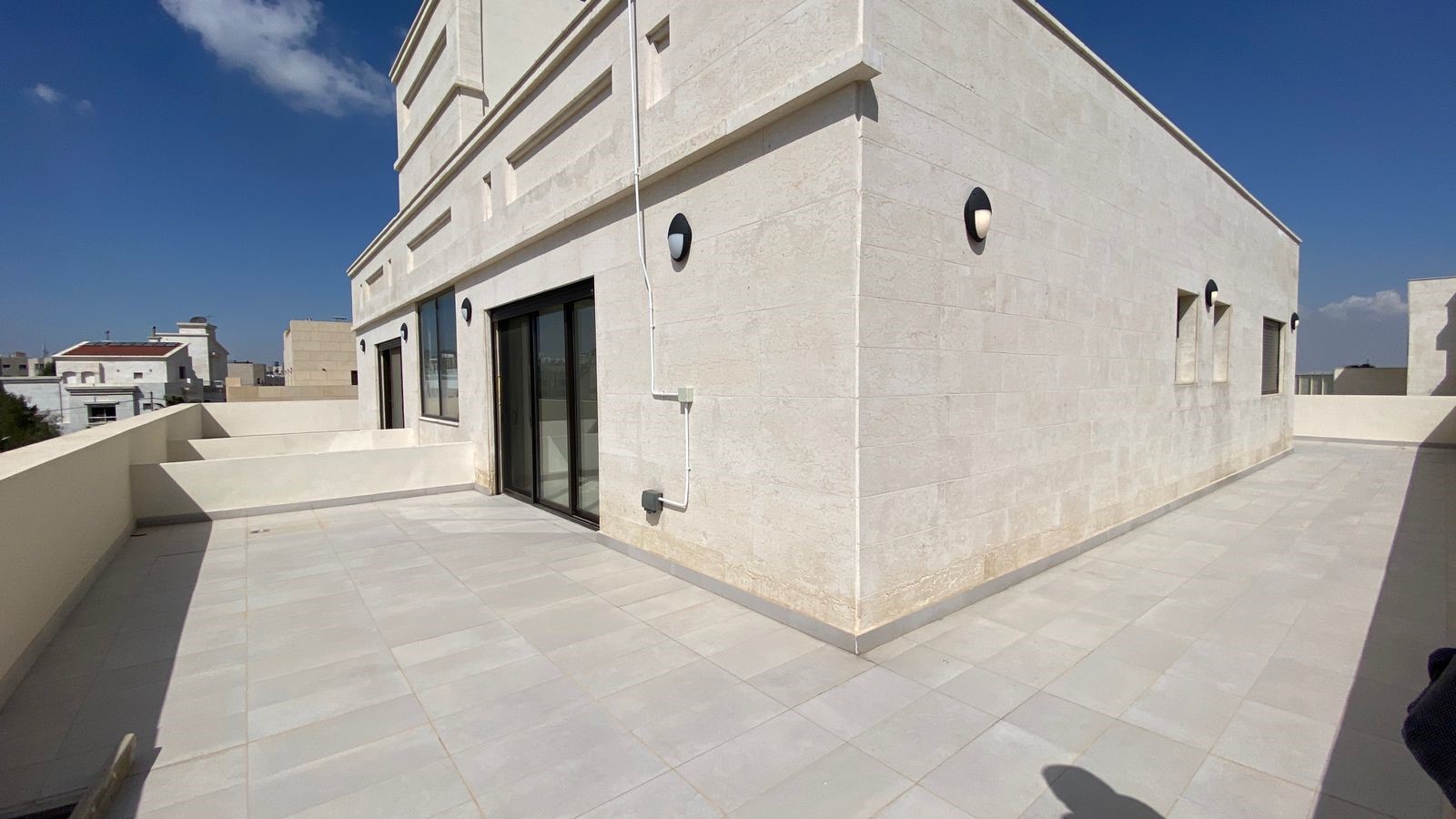 6f25e7c5-5783-4381-b713-8553f9d2c1be Rooftop terrace of a light-colored stone building with large sliding glass doors, circular wall lights, and a clear blue sky overhead.