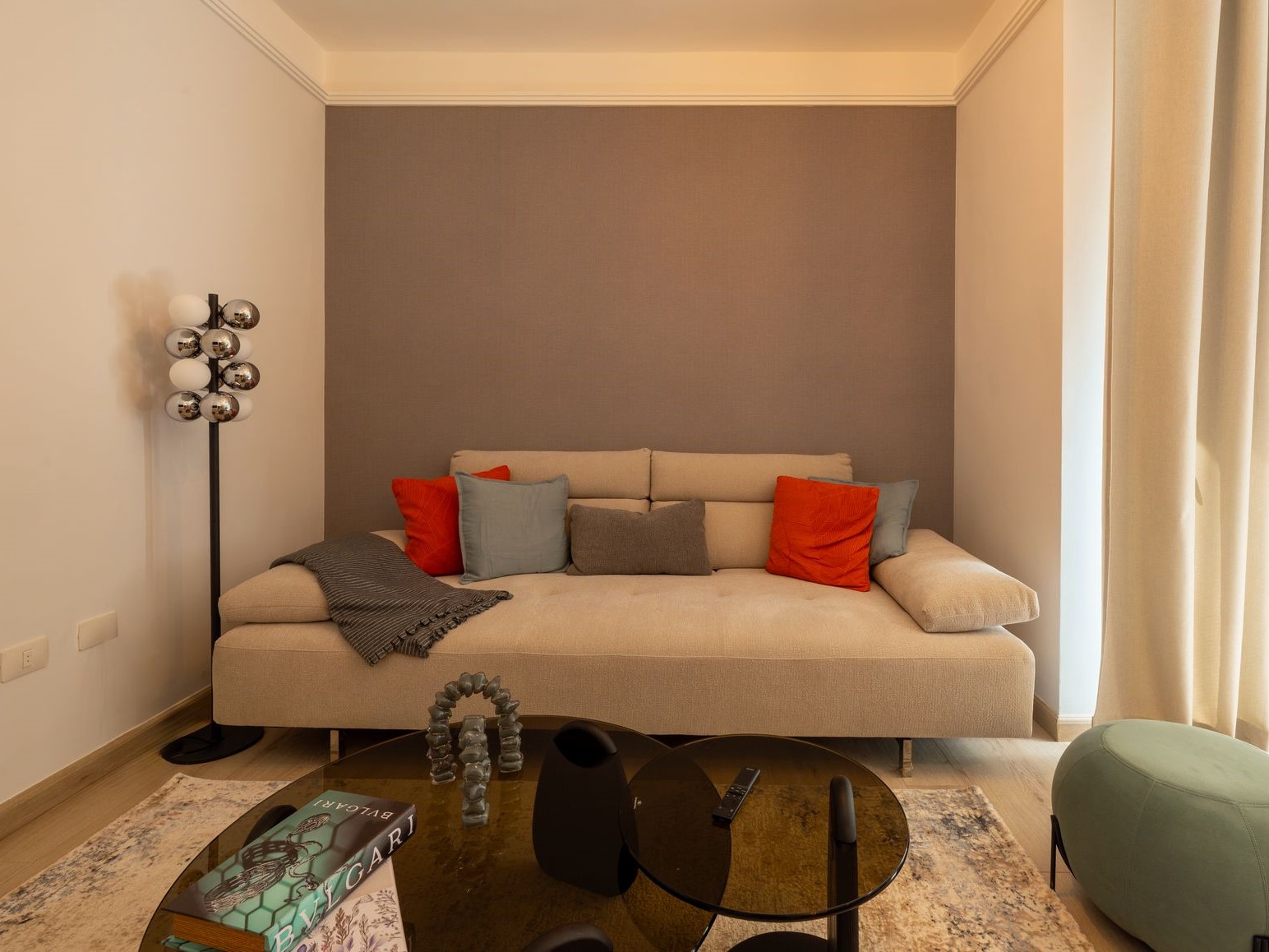 6f475278-e761-4656-9aa3-1a87b2daf55f Cozy living room with a beige sectional sofa, colorful cushions (orange, blue, gray) and a gray blanket, in front of a gray accent wall.