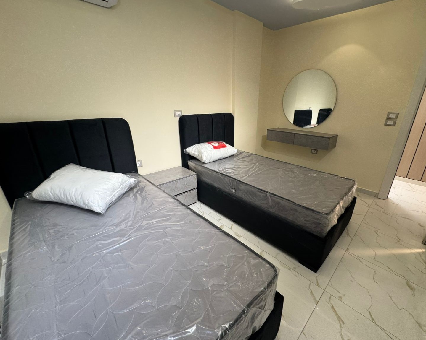 70f17835-aa6e-4a6e-92f6-8f898847b603 Two single beds with gray sheets and black upholstered headboards in a bright hotel room, separated by a small gray nightstand.