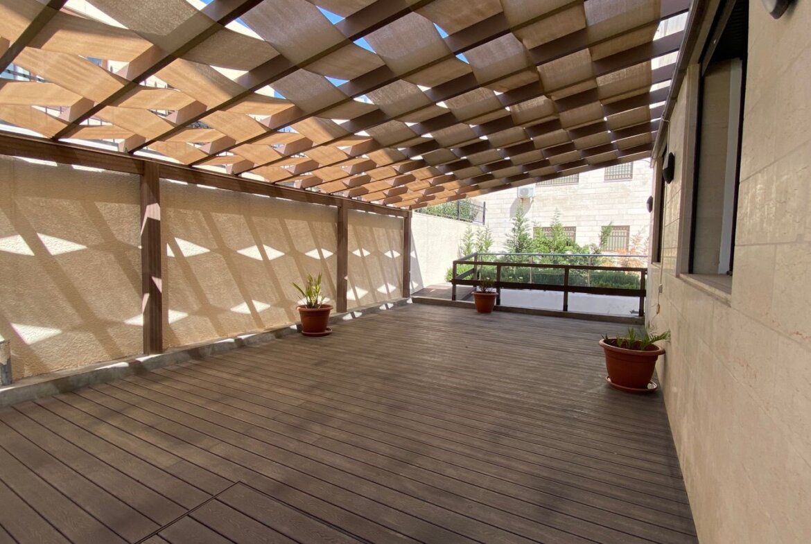 Wooden rooftop terrace with a slatted pergola casting geometric shadows on beige walls and floor. A few potted plants sit along the edges.