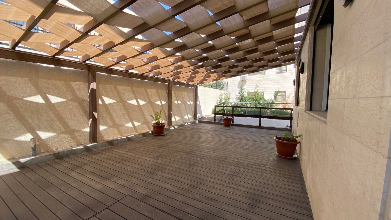 710bb583-0600-4dab-893d-e7fa68cb5e55 Wooden rooftop terrace with a slatted pergola casting geometric shadows on beige walls and floor. A few potted plants sit along the edges.