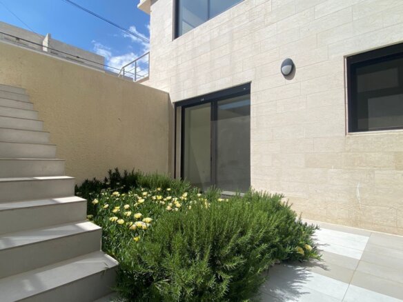 Modern building exterior with light stone walls, a wide set of cream stairs on the left, glass door and window, and a landscaped bed of yellow flowers and green shrubs in the foreground.