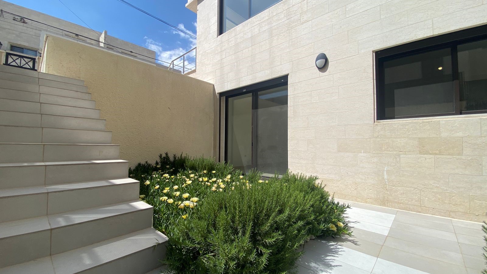 72193fdd-05cc-41fc-94fc-1c496ced9e37 Modern building exterior with light stone walls, a wide set of cream stairs on the left, glass door and window, and a landscaped bed of yellow flowers and green shrubs in the foreground.