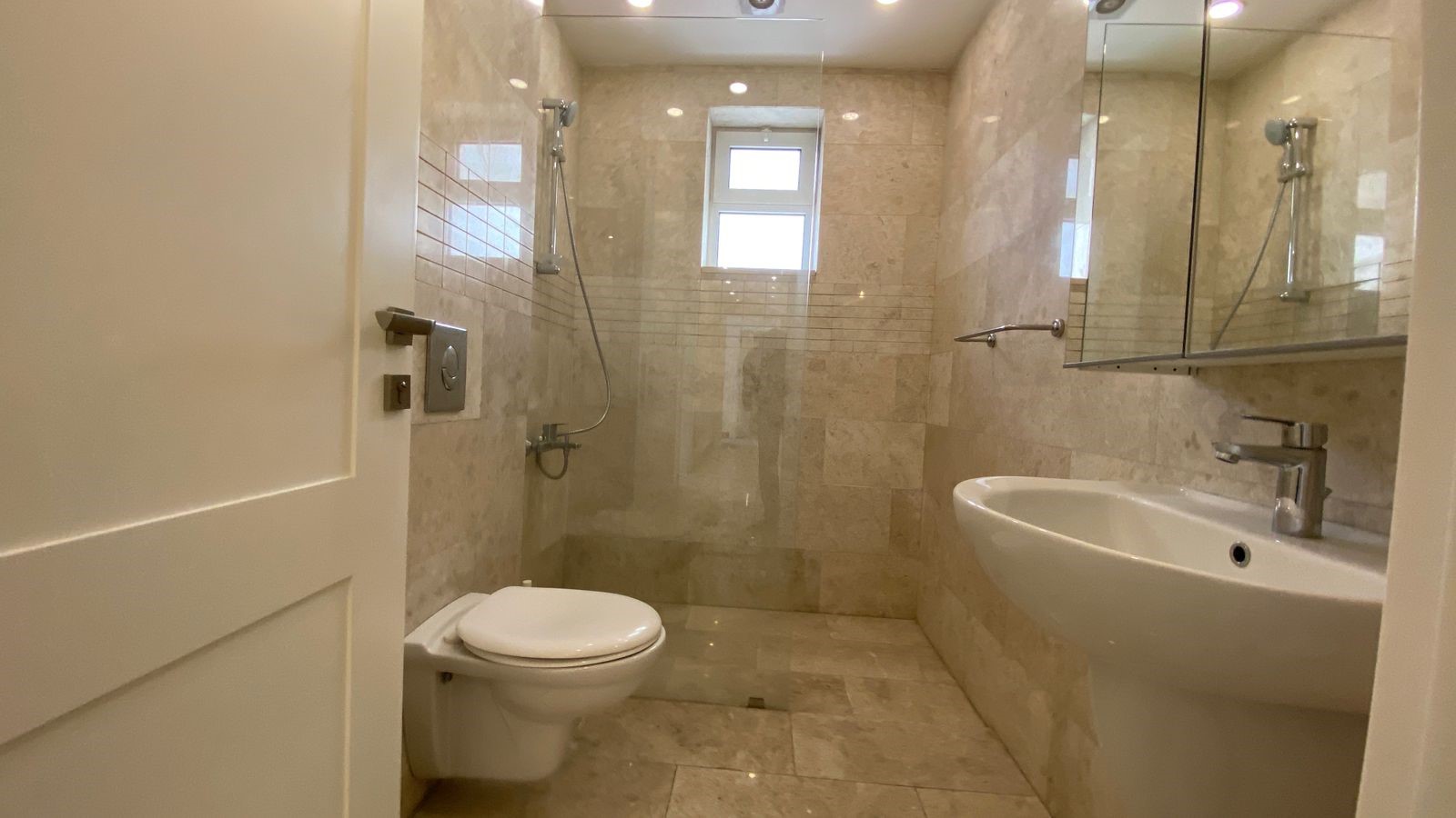 732a41fb-3b10-4f03-b56b-fa6ed44138d6 Modern beige tiled bathroom with toilet, pedestal sink, and glass shower enclosure, viewed from doorway.