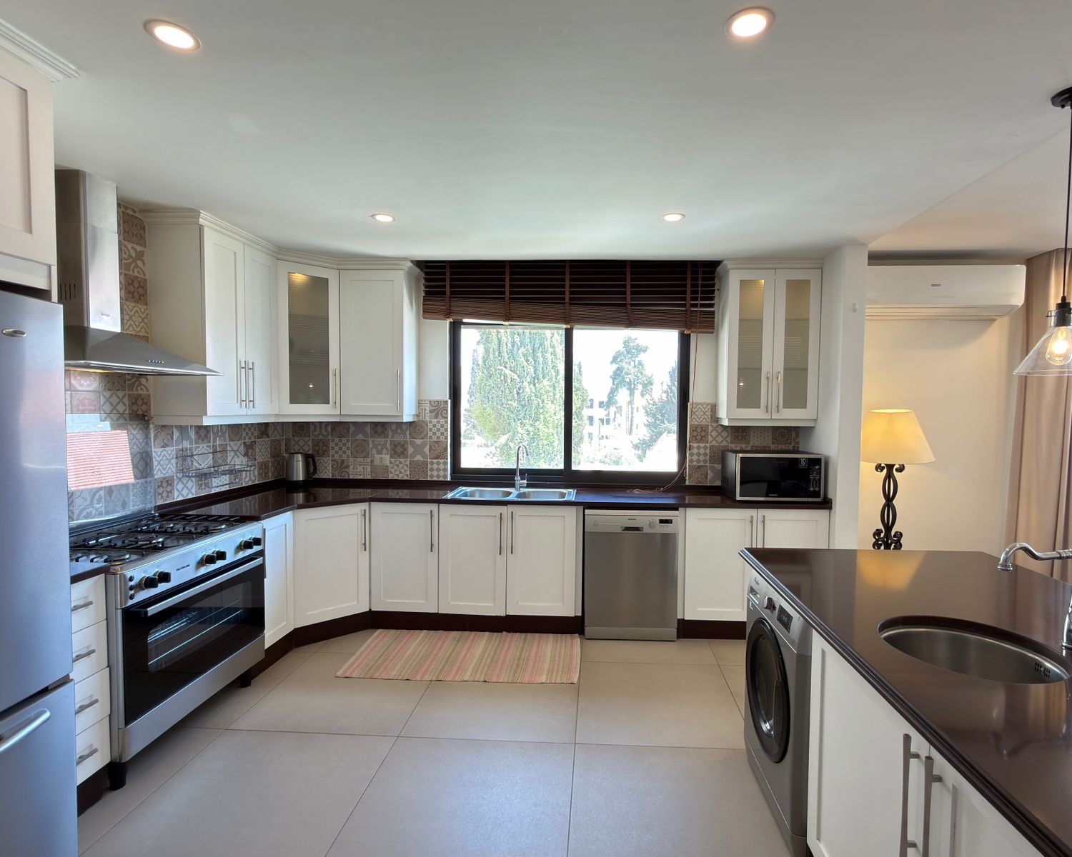 739def7c-adea-4bc9-bfe7-a3518840a82e Modern U-shaped kitchen with white cabinets, dark countertops, and a patterned tile backsplash beneath a large window over the sink