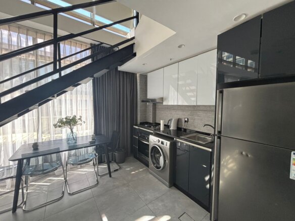 Modern kitchen under a black steel staircase with a stainless fridge, washing machine, and white upper cabinets opposite a glass dining table by large windows.