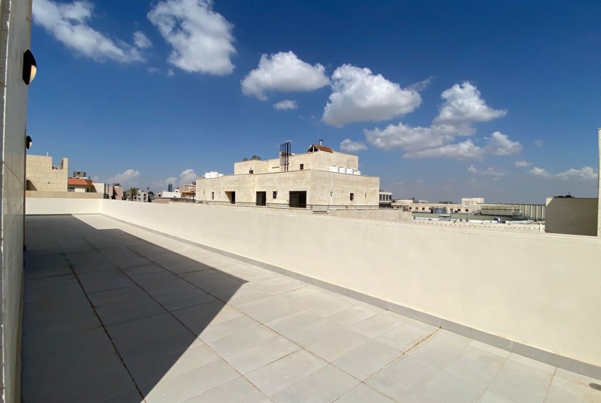 Sunlit rooftop terrace with light tiles and a low white wall, overlooking beige stone buildings against a blue sky with clouds.
