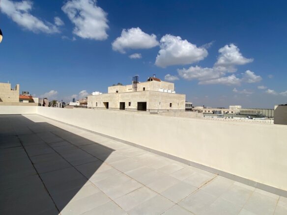 Sunlit rooftop terrace with light tiles and a low white wall, overlooking beige stone buildings against a blue sky with clouds.