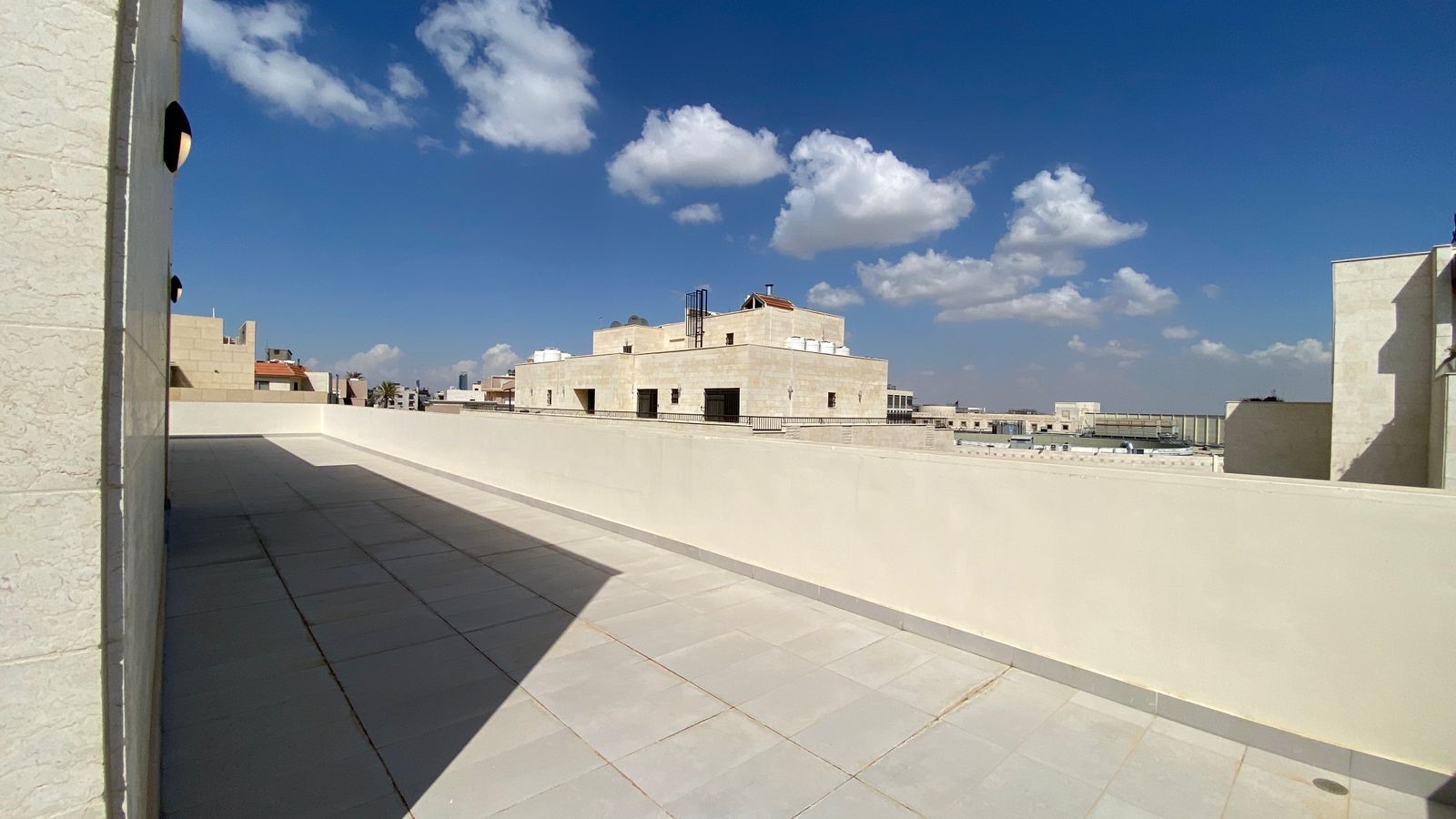73e74f4b-2530-4630-b581-a7d4edbe200b Sunlit rooftop terrace with light tiles and a low white wall, overlooking beige stone buildings against a blue sky with clouds.