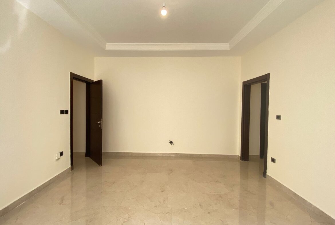 Empty beige-walled room with a tiled floor, two doorways (one left open, one right) and a single ceiling light bulb overhead.