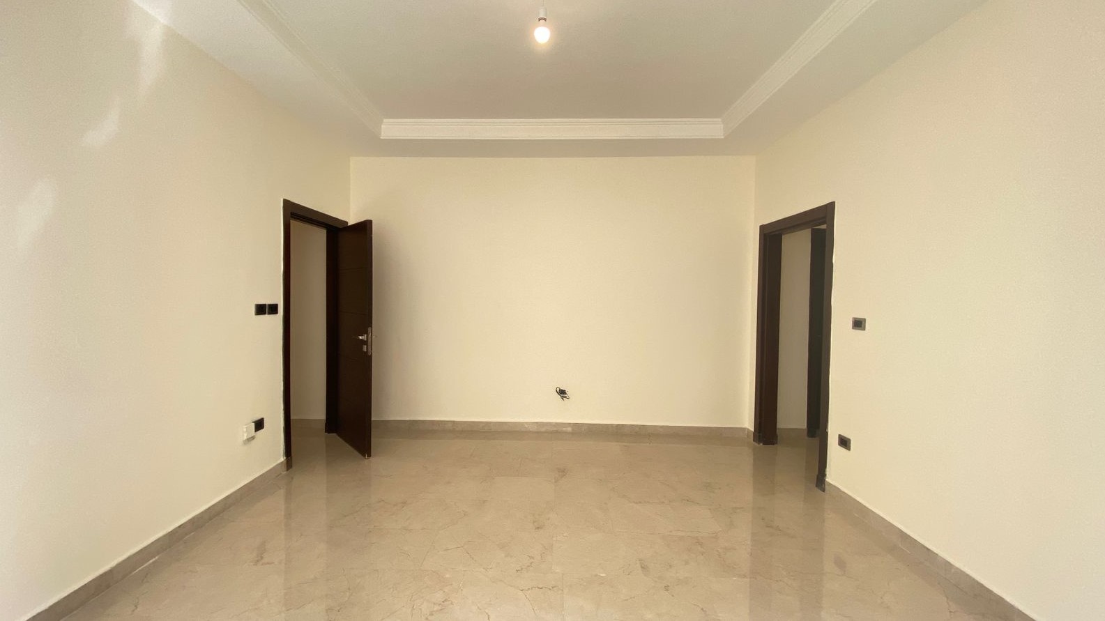 750e7945-e09f-4bc1-9208-04862855ecde Empty beige-walled room with a tiled floor, two doorways (one left open, one right) and a single ceiling light bulb overhead.