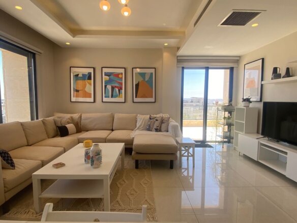 Bright living room featuring a beige sectional sofa, white coffee table, and a row of abstract art frames above. Large glass doors open to a sunny balcony.