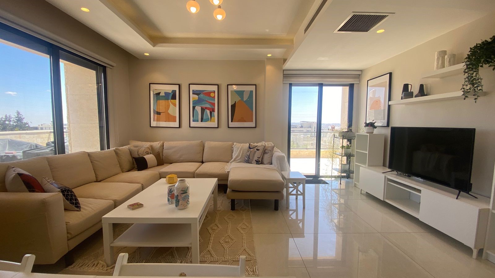 7589c60e-baba-463a-b7da-523996522b2b Bright living room featuring a beige sectional sofa, white coffee table, and a row of abstract art frames above. Large glass doors open to a sunny balcony.