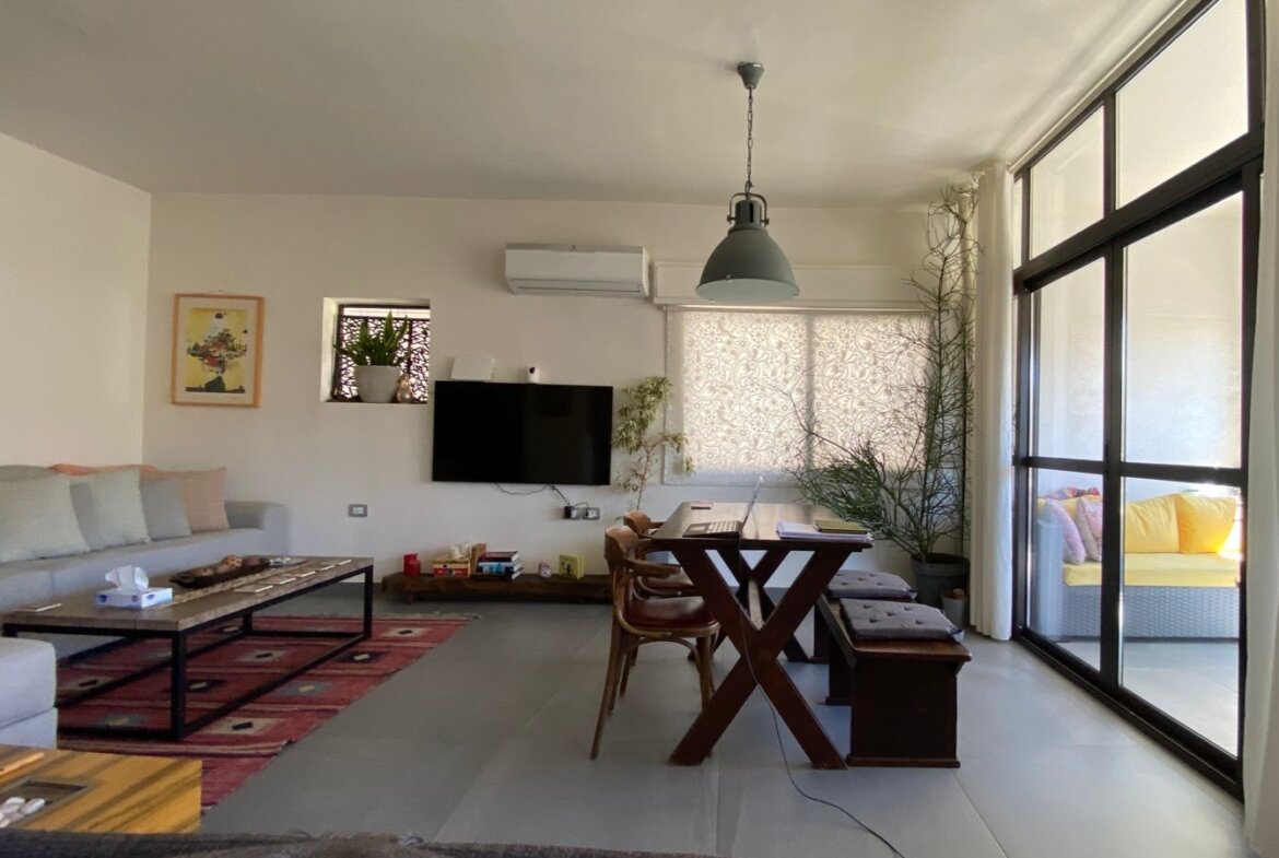 Bright open-plan living room with a white sofa, wall-mounted TV, and a wooden dining table near large glass doors to an outdoor seating area.