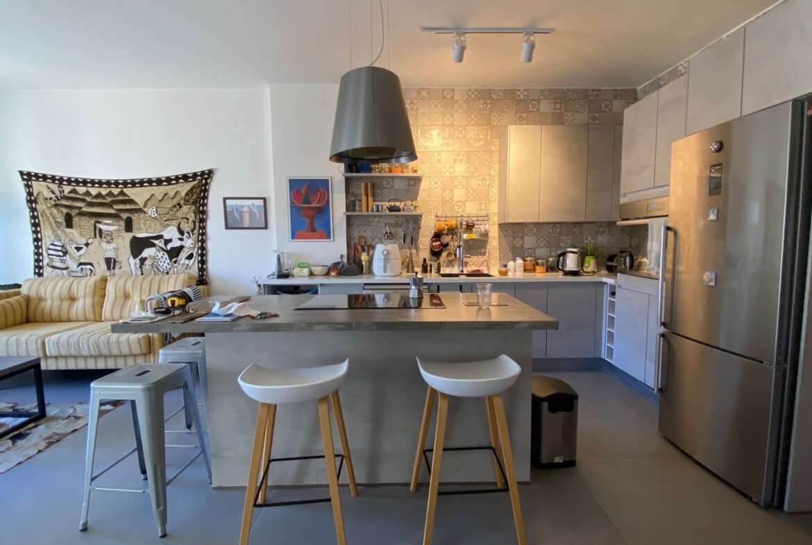Open-plan kitchen with a central island, two white bar stools, and a stainless steel fridge; living room seating and wall art visible to the left.