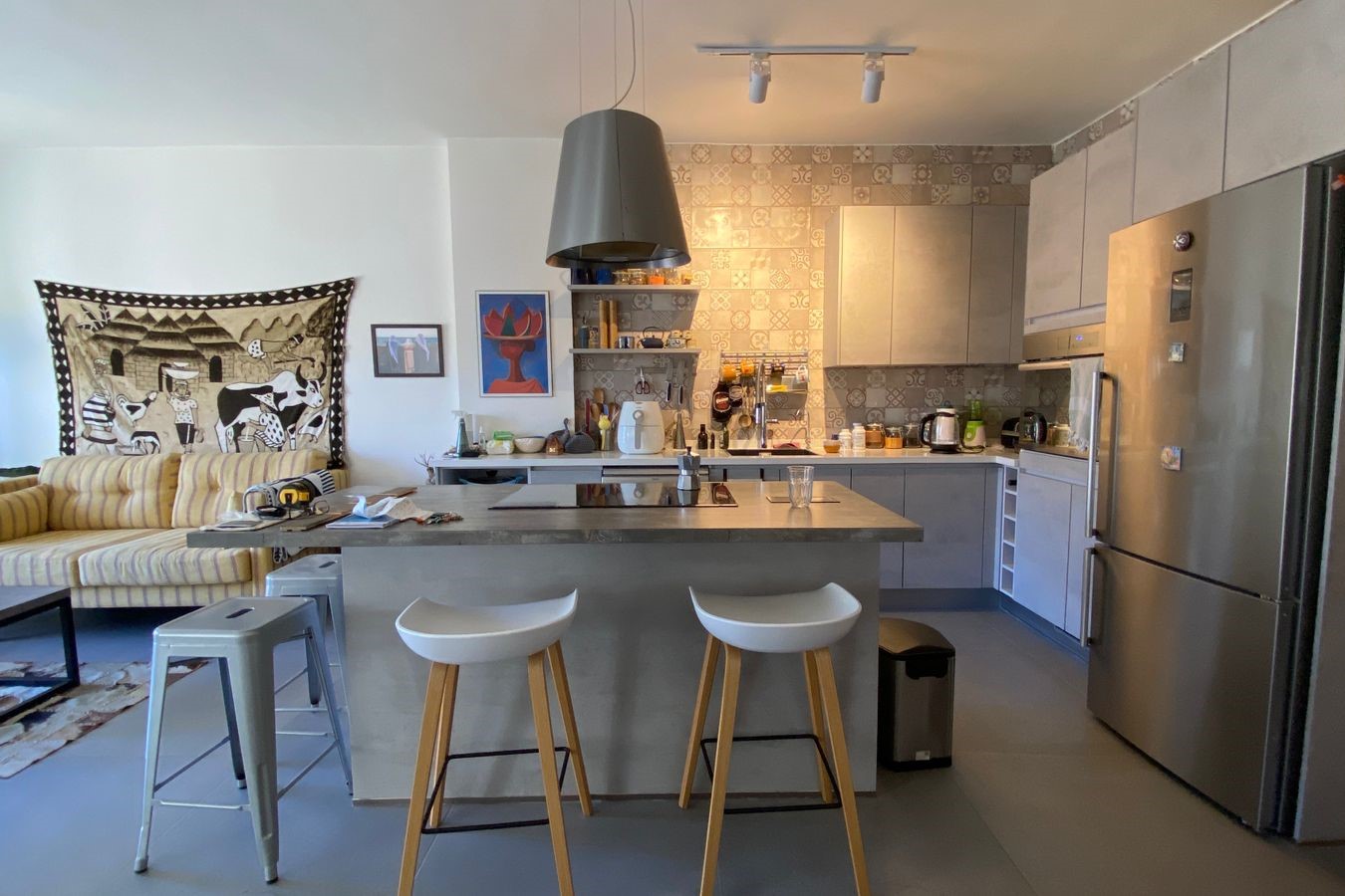 Open-plan kitchen with a central island, two white bar stools, and a stainless steel fridge; living room seating and wall art visible to the left.