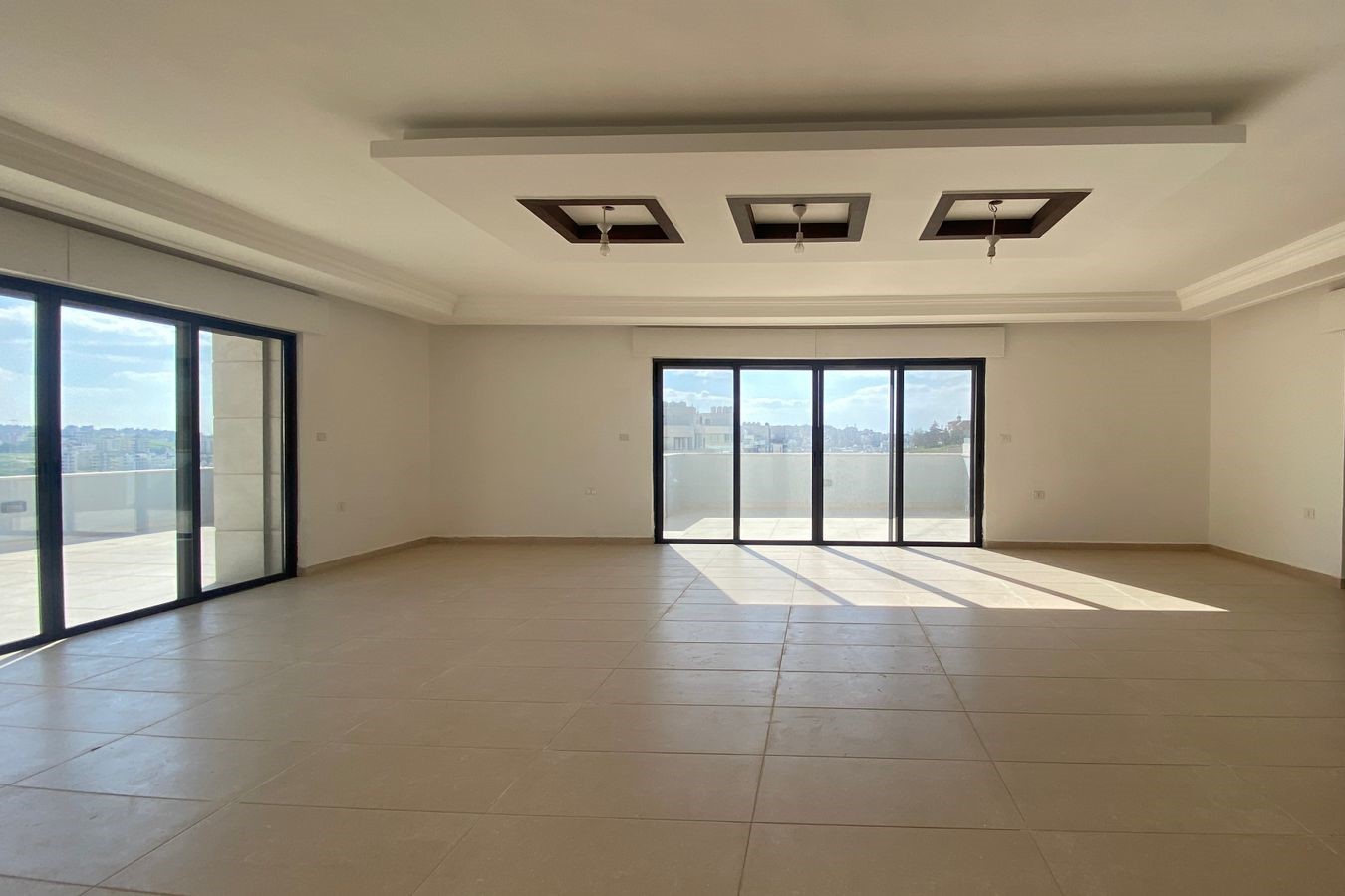 76b2fe9a-d996-4bdb-85f4-77f12cd44212 Empty living room with large glass sliding doors to a balcony, tiled floor and a recessed ceiling with hanging light bulbs.